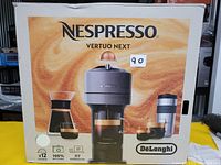 Photo of Nespresso Vertuo Next coffee machine box showing machine, carafe, travel mug, and branding.