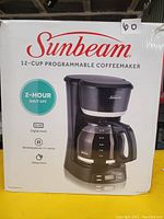 Boxed Sunbeam 12-Cup Programmable Coffeemaker showing front packaging with features listed such as 2-hour shut off, digital clock, brewing pause-to-serve, and delay brew.