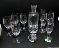 Photo showing a clear glass decanter with an air bubble in its bottom alongside 8 clear glasses: 4 wine glasses and 3 flute glasses, all arranged on a black background.