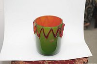 Front-facing photo of the vase showing green and orange glass layers and red zigzag decoration on white background.