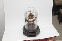 Full view of skeleton clock with visible gears under glass dome on octagonal marble pedestal base.