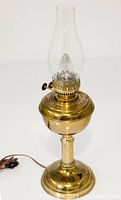 Full view of vintage brass oil lamp with tall glass chimney and electrical plug cable visible at base.