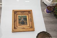 Full view of the oil painting in ornate gold frame placed on white surface with a metal tray partially visible.
