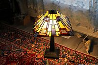 Lamp illuminated in indoor setting showing stained glass shade with yellow, blue, white, amber, and brown geometric panels, base visible on patterned rug.