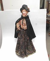 Full front view of the Dicken's style Victorian doll dressed in a black cape and patterned skirt, showing hands and facial details.