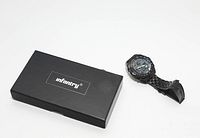 Black Infantry wrist watch next to the black Infantry branded box, showing the watch's textured strap and round dial.