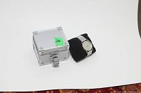 Photo showing silver wrist watch and metal storage box with green lot number sticker.