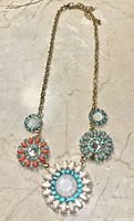 Photo of a gold-tone necklace with four circular pendants featuring turquoise-colored, coral-colored, and white beads with central iridescent stones, laid flat on a marble surface.