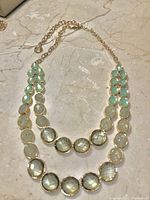 Photo of double strand blue-green faceted crystal necklace with gold-tone chain lying flat on a marble surface.