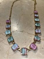 Image showing a gold chain necklace with rectangular alternating pink and blue gemstones resembling rose quartz and aquamarine, arranged symmetrically along the chain.