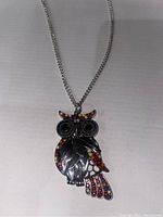 Full view of owl pendant necklace on a plain surface, showing silver-tone detailing and colorful stones on wings and tail.