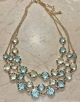 Front view of the necklace showing three rows of blue and light green round faceted stones in gold-tone settings on a gold chain.
