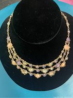 Three rows of gold-tone chain necklace featuring polished oval and round smoked topaz and lilac opal stones, displayed on a black necklace stand.