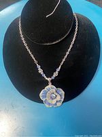 Photo of the necklace displayed on a black bust with visible flower pendant in blue and white enamel and small blue and clear beads on the chain.