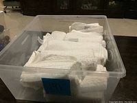 Clear plastic bin containing 5 medium white bathroom towels and 9 white face cloths, folded and stacked.