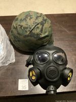 Full view of army helmet covered in digital camouflage fabric with black gas mask placed in front on a wooden surface.