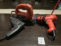 Three corded power tools on a dark tabletop: Black and Decker reversible drill, black Skil drill, and Black and Decker jigsaw.