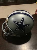 Left side view of silver Dallas Cowboys helmet with blue star logo, face mask attached, resting on dark surface.