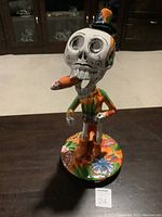 Photo of the front side of the Day of the Dead figurine showing detailed skull face with cigar and colorful outfit on round base.