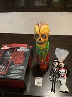 Photo showing the three colorful Day of the Dead skulls stacked vertically, the clear skull kit box, and bride and groom figurines on a dark table.