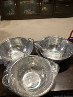 Three new stainless steel colanders in protective plastic wrap, different sizes visible