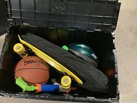 Black storage bin holding some of the items including yellow skateboard, badminton set bag, water gun, basketball, and helmet