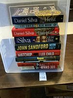Photo shows a stack of 10 books with visible titles by Daniel Silva, Lee Child, John Sandford, and John Grisham in a plastic box.