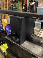 Samsung black soundbar placed on top of black subwoofer in garage with various items in the background.