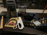 Photo showing full lot of various tools including stapler, screwdriver sets, mallet, hole saw kit, glue gun, and flexible drill extension, arranged on dark wooden table.