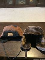 Two winter hats on a dark wooden surface including a brown knit hat with black shearling lining and a brown faux fur-lined hat.