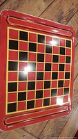 Full view of the metal checkers board showing red border, black and red squares with yellow grid, and recessed trays on opposite ends.