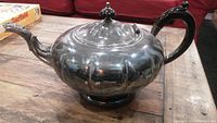 Side view of the silver plated teapot showing bulbous fluted body, ornate spout, and handle with dark grip, sitting on wooden surface.