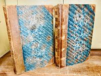 Two hardcover volumes standing upright, showing front covers with light brown leather spine and corners, and blue marbled paper covering rest of the boards. Both show wear to spine and covers.