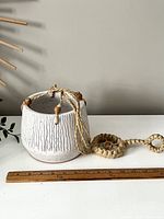 White textured pottery hanging planter suspended by jute rope with wooden beads, shown on a white surface with a wooden ruler for scale.