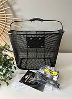 Front and side view of black metal mesh bike basket showing rectangular shape, metal mesh weave, plastic handle grip, and mounting points with included hardware in plastic bags, placed on white surface with instruction sheets beneath.