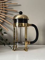 Bodum French press coffee maker upright on surface showing gold metal frame, glass beaker, black handle, and lid with black knob.