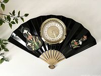 Front view of black lacquered fan clock with central round clock face and floral decoration on each side.