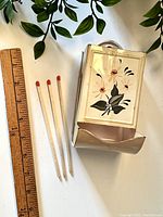 Front view of vintage tin matches container with flower painting next to ruler and three matches lying beside.