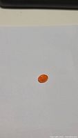 Photo showing small oval-shaped bright orange gemstone on white background, highlighting color and size.