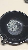 Black bowl containing numerous tiny off-white, white, and light teal zircon gemstones, each about 1mm in diameter.