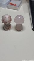 Photo of two hand-carved natural gemstone mushrooms; left mushroom likely jasper or agate with reddish-brown veining, right mushroom rose quartz pale pink, both standing on flat surface.
