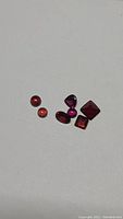 Photo shows seven loose red garnet gemstones of various shapes and cuts on a light background.