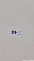 Pair of small round diamond-cut tanzanite gemstones with purple-blue coloring on plain background.