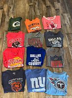 Full set of 12 graphic crew neck women's t-shirts, displaying sports and team logos in various colors and sizes, mostly large with one XL.