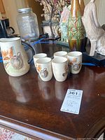 Ceramic pitcher and five matching cups showing wine and cheese themed artwork with visible crazing texture on surface.