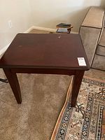 Photo showing solid wood end table with dark finish. Top surface shows wear with light scratches and a larger spot worn through finish. Table sits on carpet near sofa and decorative rug.