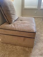 Full side angle view of beige corduroy fabric ottoman featuring dark faux leather piping, tufted top cushion, and matching pillow placed on carpet.