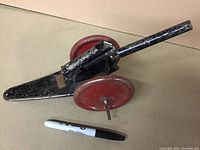 Full side view of antique metal toy cannon with red wheels placed next to a Sharpie pen for size reference