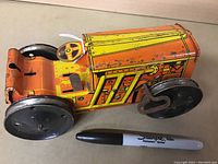 Side view of Marx wind-up tin toy tractor showing orange and yellow lithographed body, black wheels, and winding key with Sharpie marker for size reference