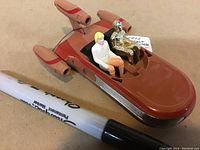 Front side of the Landspeeder toy with Luke Skywalker and C-3PO seated inside next to a marker for scale.